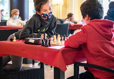 girl and boy playing chess wearing masks red table